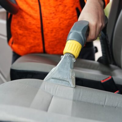 Osoba w pomarańczowej kurtce wykonuje czyszczenie tapicerki w samochodzie za pomocą specjalnego odkurzacza.